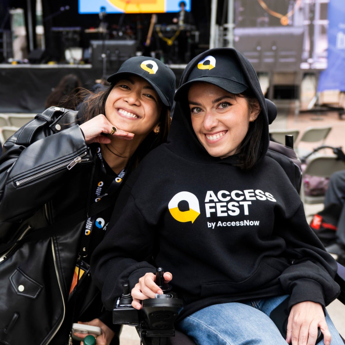 Two women smiling together at an outdoor event with a stage and 'Access Fest' branding. They are wearing black baseball caps with the yellow and white arrow AccessFest logo