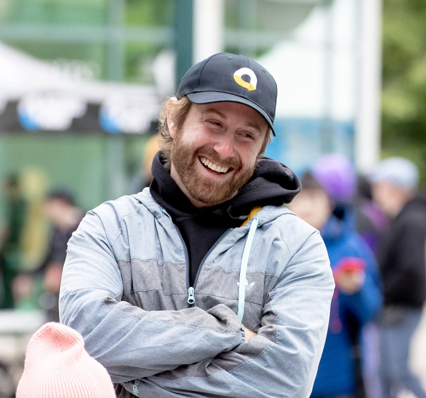 A man with red hair wearing a gray jacket and black cap with a yellow and white logo on it, stands outdoors, laughing. Blurred people and event tents are visible in the background.