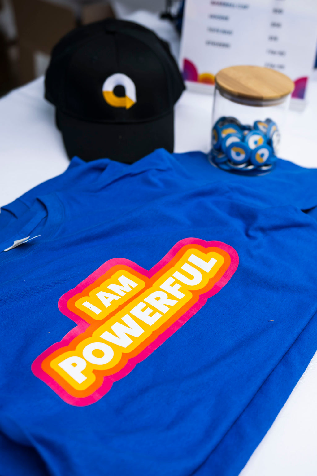 Blue t-shirt with 'I AM POWERFUL' text on a white surface with a black cap and jar of AccessFest logo buttons in the background.