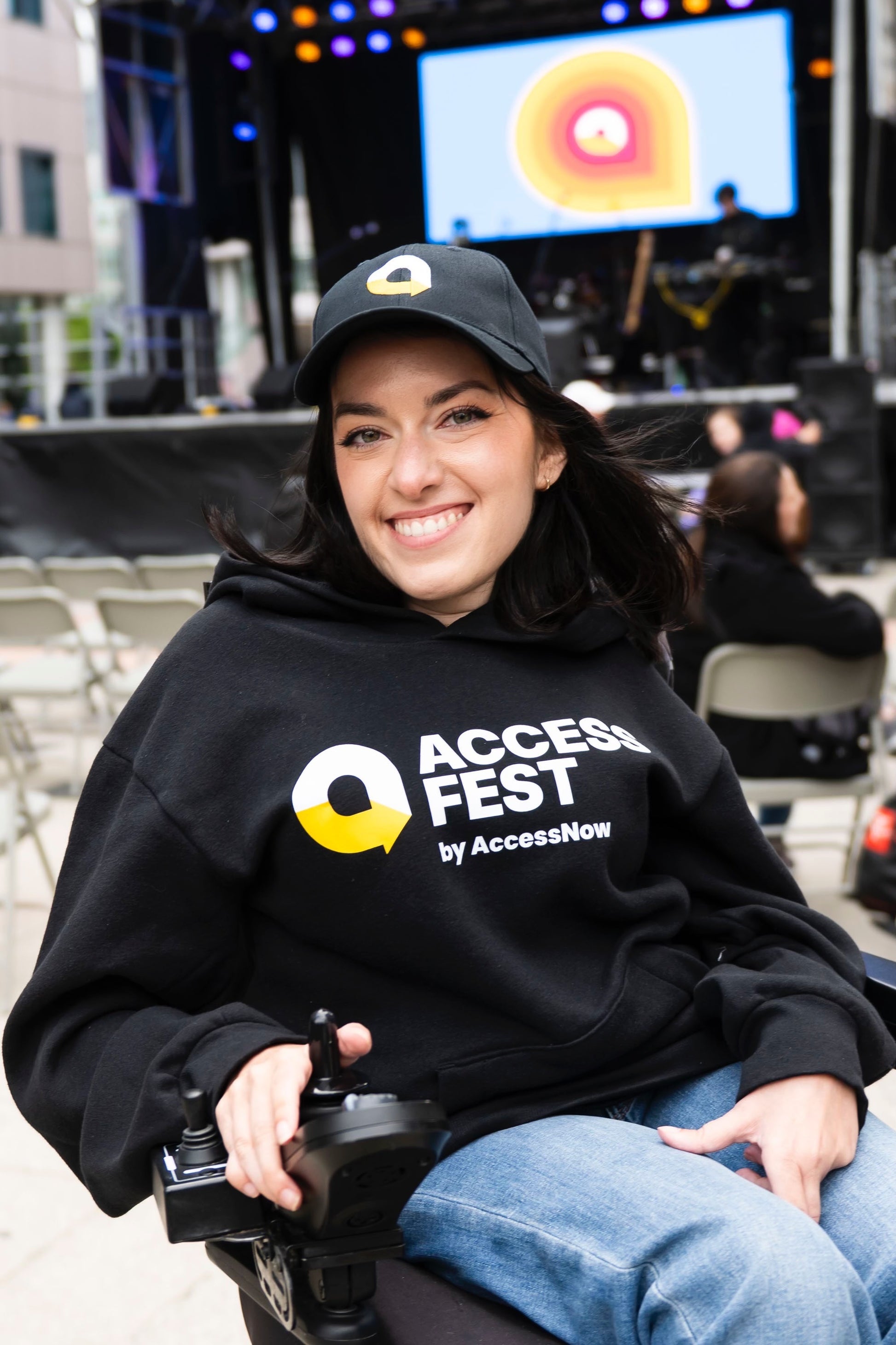 Maayan Ziv, a woman with long dark hair sits in a power wheelchair, smiling at the camera. She wears a black AccessFest hoodie and matching black cap with the yellow and white AccessFest icon. The background shows a stage with colorful lights and a large screen displaying the AccessFest logo, with people seated in folding chairs at the outdoor event.