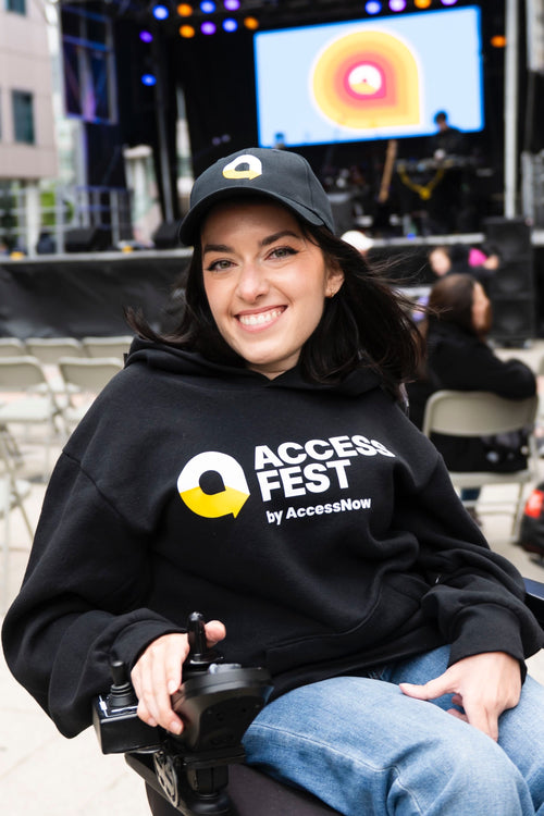 Maayan Ziv, a woman with long dark hair sits in a power wheelchair, smiling at the camera. She wears a black AccessFest hoodie and matching black cap with the yellow and white AccessFest icon. The background shows a stage with colorful lights and a large screen displaying the AccessFest logo, with people seated in folding chairs at the outdoor event.
