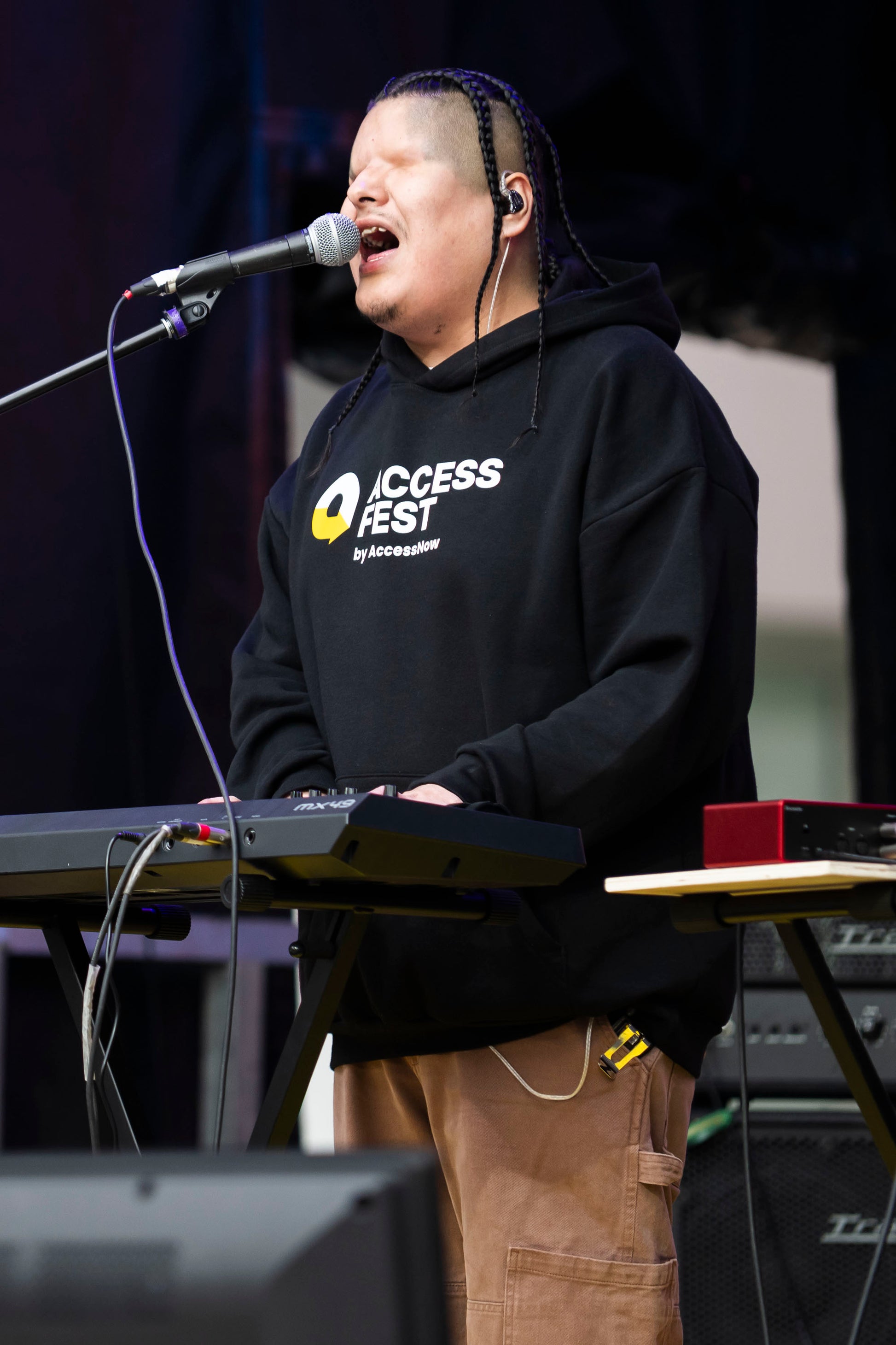 MattMac, an Indigenous blind man, wearing a black hoodie with the AccessFest logo, singing into a microphone and playing a keyboard on stage.