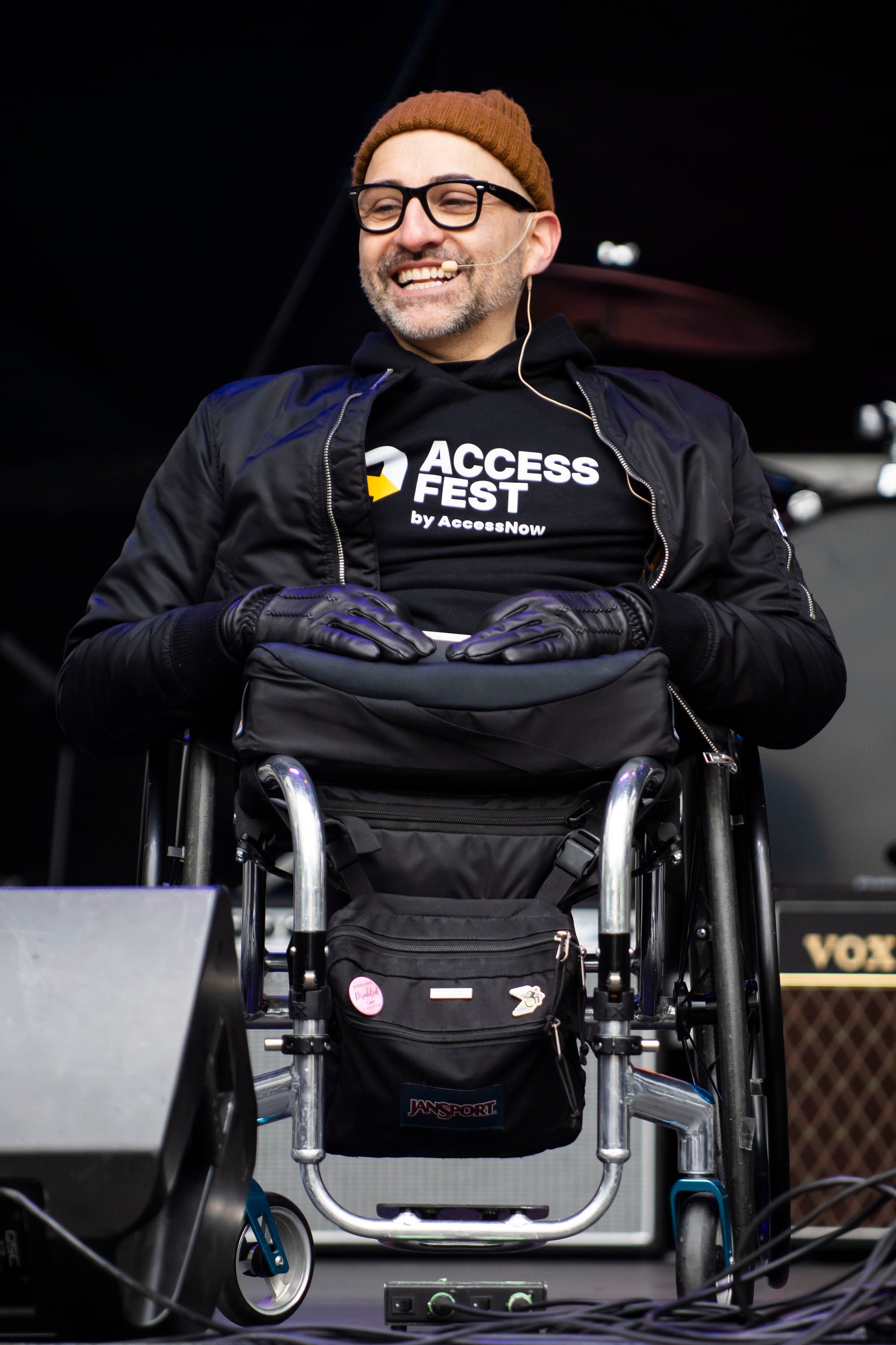 Spencer West, a white man with a beard, wearing glasses, a brown beanie, and black gloves, smiles while seated in a wheelchair on stage. He wears a black AccessFest by AccessNow hoodie and bomber jacket, with a microphone headset visible. Stage equipment and instruments are visible in the background.

