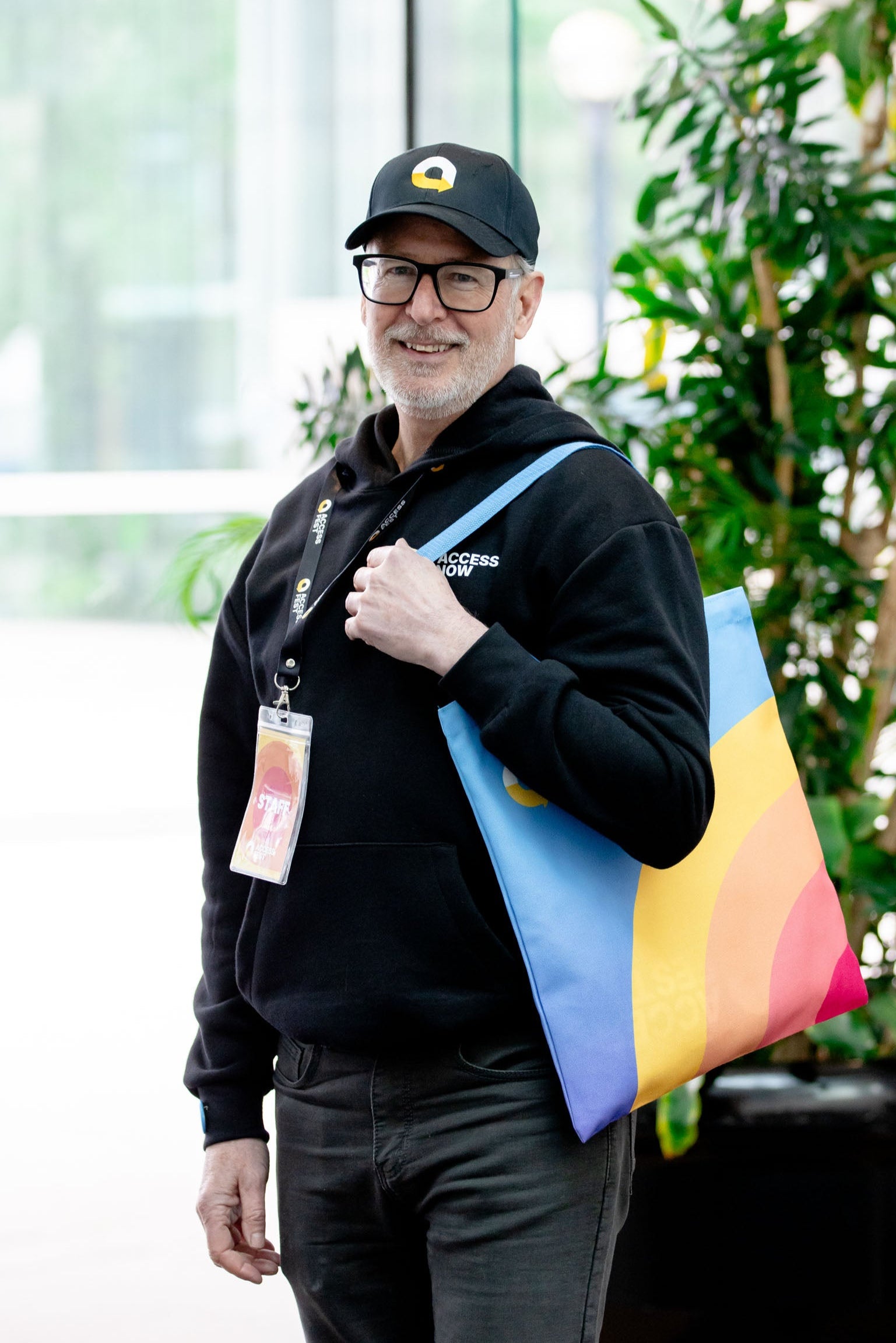 A white middle-aged man wearing a black hoodie and black cap, holding a colorful tote bag. He stands in front of a blurred indoor background.

